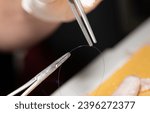Small photo of Close-up of a surgical clamp and forceps gripping surgical suture material. In the background, a blurred hand in a glove and a blurred surgical specimen.