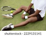 Close-up of the hands of African-looking tennis player sitting on the grass of tennis court. Next to him lies tennis racket and ball. Guy is wearing white T-shirt, black shorts and sneakers.