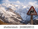 Small photo of The crisp air of an alpine road is marked by a cautionary traffic sign, warning of the curving path ahead. Behind, the grandeur of snow-dusted mountains looms, signifying the beauty and perils of moun