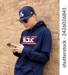 Small photo of Chicago, Illinois - February 15, 2024: A focused young man, clad in a White Sox cap and hoodie, is engrossed in his smartphone against a brick wall backdrop