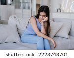 Small photo of Disappointed young beautiful Hispanic girl sitting on cozy sofa with cushions leans on hand looks down in upset mood. Troubles, financial problems. Perplexed female at home. Failure, lack of energy.