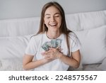Small photo of Excited blonde caucasian girl broad smiling eyes closed presses banknotes to chest, got salary, sits on bed. Happy Swedish young woman received money, celebrates. Prize, lottery, winner. Wage. Cash