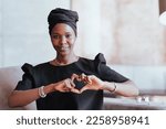 Cheerful African woman In black turban and black dress sitting on cozy sofa makes heart gesture by hands looks at camera broad smiles. Happy Brazilian female demonstrates her emotions. Love concept.
