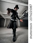 Small photo of From below full body of emotionless bearded african american male with accessory on hands in extravagant black outfit and tall hat dancing in garage near brick wall