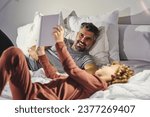 Cheerful man and boy in sleepwear having fun while lying on soft bed and reading interesting book together in cozy bedroom at home