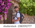 Small photo of Side view of emotionless mature man in cap standing near old building with stone brick wall and looking at camera while travelling on summer vacation
