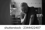 Small photo of Senior man suffering from depression at home alone leaning by kitchen sink in dramatic black and white, monochromatic scene of person in despair succumbed by hardship