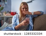 Heartbroken blond girl holding pink smartphone while sitting on couch. She looking at phone display with unhappy expression and covering mouth with hand as if going to cry