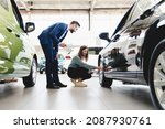 Small photo of Beautiful young caucasian female client customer choosing new car, trying checking its options, tire, wheels while male shop assistant helping her to choose it at dealer auto shop