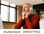Relaxed mature middle-aged man businessman freelancer listening to the music, podcast, e-book, radio programme, playlist, musical track in headphones while working remotely on laptop at home