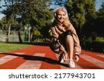 Small photo of Caucasian mature female runner athlete tying training shoes while jogging on the stadium in public park in the morning. Active healthy sporty lifestyle.