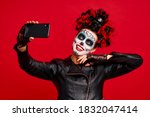 Girl with sugar skull makeup with a wreath of flowers on her head and skull, wearth lace gloves and leather jacket,making selfi isolated on red background. concept of Halloween or La Calavera Catrina.
