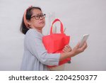 An Asian woman holding shopping bags has difficulty looking at her cell phone.