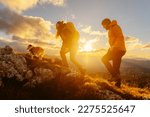 Small photo of Two mountaineers against the light, in silhouette, with the sun in the background, ascending a mountain peak with their dog at sunset. Traveling with a pet. Sport and physical activity outdoors.