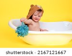Small photo of Unhappy baby toddler boy is sitting in a white bathtub on a studio yellow background. Squeamish child at the age of one year
