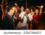 A cheerful group of multiracial people is dancing, drinking beer, and having a good time at the outdoor nighttime event.
