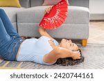 Small photo of A relaxed woman lies on the floor with a bright red fan, finding respite from the heat inside her home.