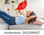 Small photo of A serene woman lies back on a sofa with a red floral fan, seeking respite from the heat.