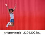 smiling african american young girl in blue shorts and white t-shirt jumping with open arms over red background