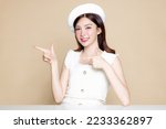 Cute Asian woman with perfect skin pointing to copy space. Pretty girl model wearing white beret and natural makeup on beige background. Cosmetology, beauty and spa, wellness, Plastic surgery.