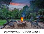 Roaring fire in a stone firepit with wood logs and surrounded by trees at dusk in a backyard