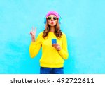 Fashion pretty sweet carefree woman listening music in headphones with smartphone wearing a colorful pink hat yellow sweater sunglasses over blue background