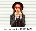 Fashionable portrait of stylish young woman covering her eyes with red heart shaped lollipop blowing her lips sending sweet kiss wearing black round hat, leather biker rock jacket on white background