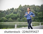 Small photo of Happy millennial Asian woman in active sportswear listening to music while enjoys running at the park in the morning. lifestyle concept