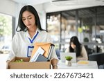 Small photo of Sad and upset millennial Asian female office worker holding a cardboard box with her belongings, walking out the company, being fired from her boss.