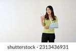 Small photo of A happy young Asian woman with a broken arm in an arm sling is looking at her smartphone screen while standing against an isolated white background.