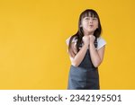 Small photo of A happy and adorable little Asian girl in a cute jeans dress is standing against an isolated yellow background in a cute pose. surprised, saying wow, excited, amazed, can't wait for the good news