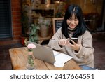 A positive, beautiful Asian woman in a cosy sweater is responding to messages on her smartphone while working remotely at a coffee shop in the city. People, wireless technology, and city life concepts