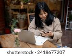 A positive, beautiful Asian woman in a cosy sweater is responding to messages on her smartphone while working remotely at a coffee shop in the city. People, wireless technology, and city life concepts