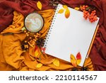 Autumn home cozy composition with yellow and burgundy blankets, cup of coffee, red and yellow leaves and notebook with copy space. Fall season template for feminine blog social media.