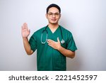 Small photo of Smiling honest asian male nurse or doctor, intern in scrubs giving pledge, raising one arm and hold hand on heart while promise, oath to patient over white background
