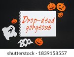 Small photo of Happy halloween holiday concept. Notepad with text Drop-dead gorgeous on black background with bats, pumpkins and ghosts