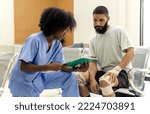Small photo of African black nurse woman asking injured informations from young Caucasian patient man in hospital, female afro hair nurse in blue hospital uniform checking beard man patient about injured wound