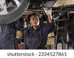 Black car mechanic woman in work cloth jumpsuit working underneath car in auto repair shop