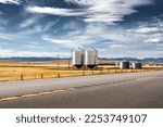 Small photo of Pair of steel grain silos standing next to a divided highway along harvested fields and distant Rocky Mountains at background near Longview Alberta Canada.