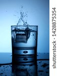 Small photo of Single glass of water ice cube trow droplets small splash fresh cold blue light liquid reflection