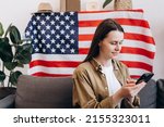 Beautiful smiling young woman sitting on cozy sofa at home with USA flag, cute happy caucasian female 20s old years with smartphone looking Independence Day celebration. Patriotic US holiday concept
