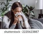 Small photo of Close up of sick woman sitting on sofa freezing blowing running nose got fever caught cold sneezing in tissue, ill brunette girl covered with blanket, having influenza symptoms coughing at home