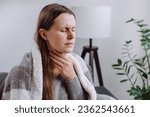 Small photo of Symptoms from flu season in winter concept. Close-up of unhealthy sad young woman covered blanket sitting on grey couch at home suffering from sore throat, angina, hard to swallow, voice loss