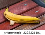 Small photo of A Closeup of The Remains of a Ripe Banana Forgotten by an Uneducated Park Visitor on a Red Bench