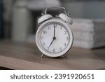 Small photo of Close-up of a round white alarm clock on a table in the bedroom. The hands on the clock show seven o'clock in the morning, time to get up. Retro alarm clock on the table, vintage tone. space for text