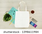 Creative, trendy, artistic eco, tote, cotton bag mock up. Mockup with earphones, a pencil,cup,paints,crayons, tassels and home plant on light wooden table, top view. Your design here, on  textile bag.