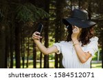 Beautiful curly-haired girl with a periphery hat and glasses selfies in the park in the spring and enjoys the sunny weather