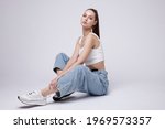 High fashion photo of a beautiful elegant young woman in a pretty white top and sneakers, blue denim jeans posing over white, soft gray background. Studio Shot. The model is sitting