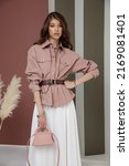 Small photo of High fashion photo of a beautiful elegant young asian woman in pretty rosy brown shirt, belt, necklace, bracelets, white cream long skirt, handbag. Green striped Corrugated Wall, dry branches.