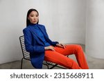 High fashion photo of a beautiful elegant young asian woman in pretty blue jacket, blazer, orange pants, trousers. White textured rounded wall. Model sits on chair